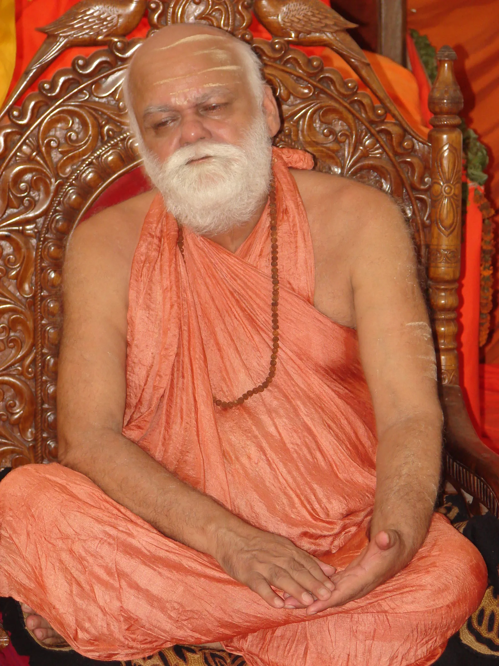 Full-length portrait of Nishchalananda Saraswati seated cross-legged on an ornately carved wooden throne, wearing saffron robes and a rudraksha mala