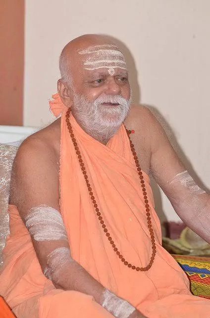 Nishchalananda Saraswati seated and smiling warmly, a rudraksha mala around his neck and sacred ash on his arms