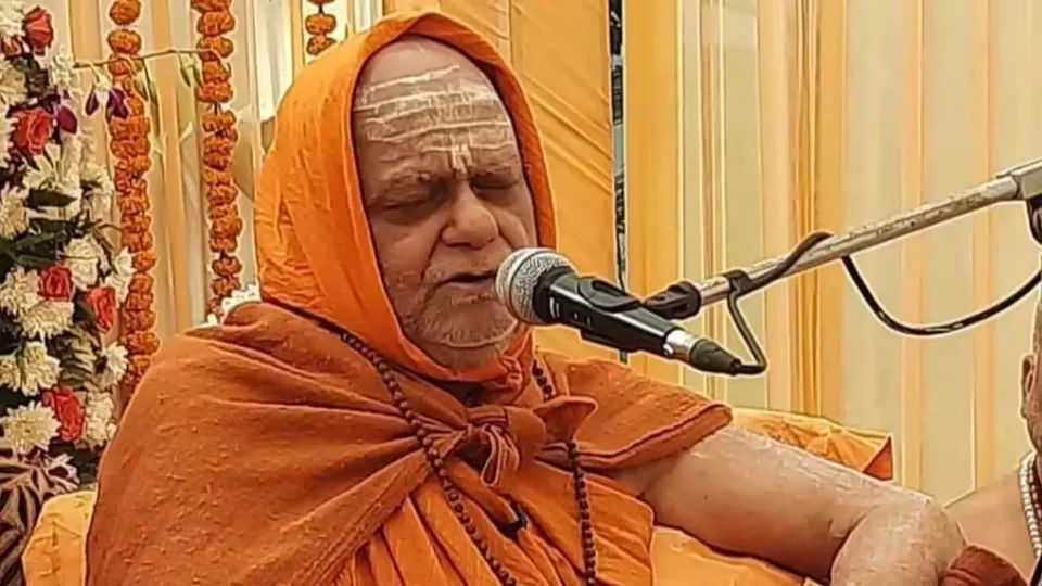 Nishchalananda Saraswati, head covered with a saffron cloth, speaking into a microphone during a discourse, with floral decorations behind him