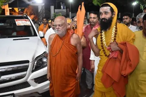 Nishchalananda Saraswati standing beside a vehicle at night, accompanied by a garlanded monk in saffron and a gathering crowd