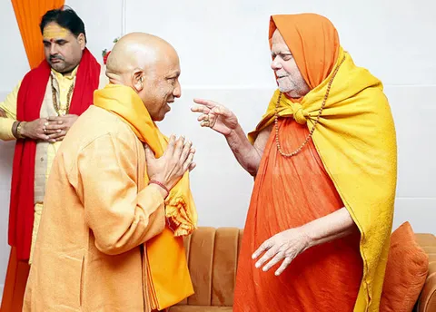 Nishchalananda Saraswati, wrapped in a yellow shawl and saffron head covering, gesturing in conversation with a visitor who listens with folded hands
