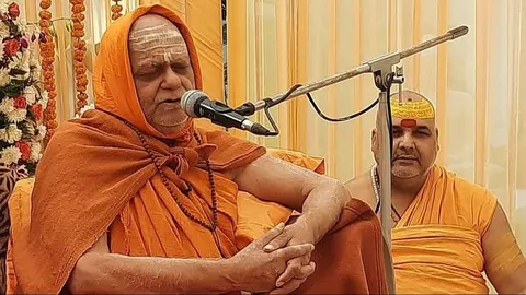 Nishchalananda Saraswati with head covered, speaking into a microphone, an attendant with a tilak seated behind him