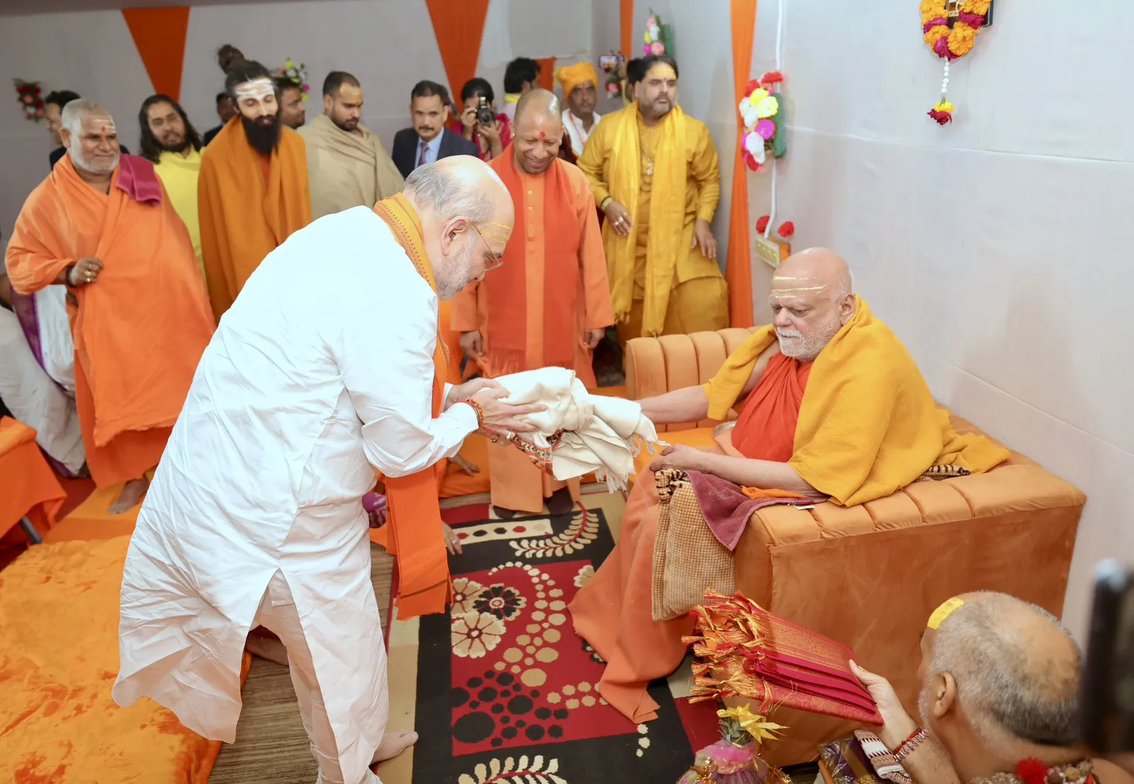 Nishchalananda Saraswati seated on a saffron-draped sofa while a visitor in white bows and offers a shawl, with monks and attendants standing behind