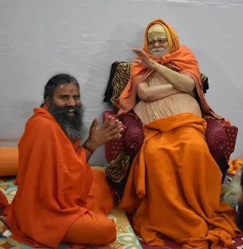 Nishchalananda Saraswati seated on a raised seat, gesturing while a bearded monk in saffron sits beside him with palms joined