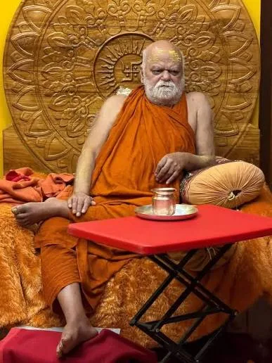 Nishchalananda Saraswati seated before a large carved golden backdrop bearing a wheel motif, a small red table with a copper vessel before him