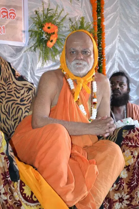 Nishchalananda Saraswati seated on a dais at a gathering, garlanded, with an Odia-language banner and floral decoration behind him