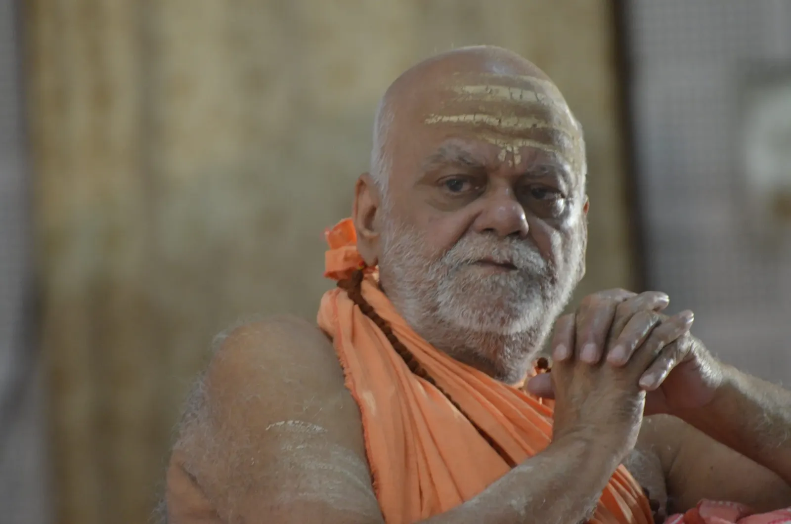Jagadguru Shankaracharya Nishchalananda Saraswati seated in saffron robes with hands clasped, a white tilak on his forehead