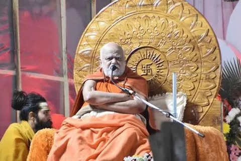 Nishchalananda Saraswati seated on a ceremonial seat before a large carved golden backdrop, addressing an assembly through a microphone