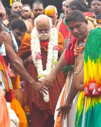 Nishchalananda Saraswati, heavily garlanded with white and pink flowers, being escorted through a crowd of devotees