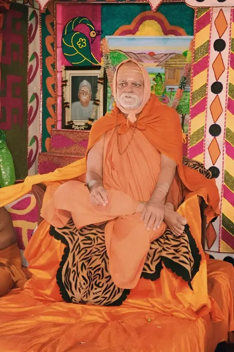 Nishchalananda Saraswati seated cross-legged on a decorated dais, a framed portrait placed on the richly painted backdrop behind him