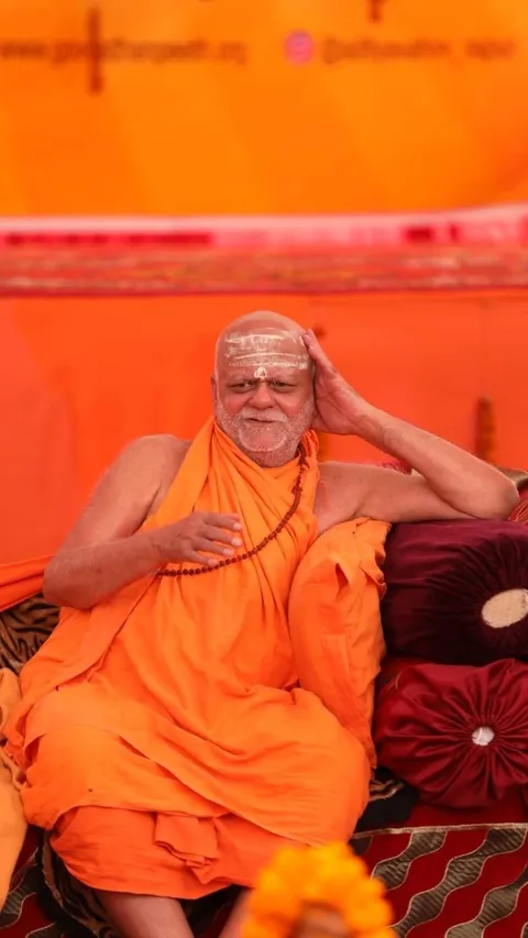 Nishchalananda Saraswati seated on cushions in saffron robes, one hand resting against his head in a reflective posture