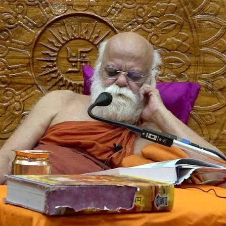 Nishchalananda Saraswati wearing spectacles, reading from a large, well-worn book placed on a cushion before him, a microphone nearby