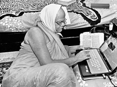 Black-and-white photograph of Nishchalananda Saraswati, a cloth draped over his head, typing on a laptop with an open book beside him