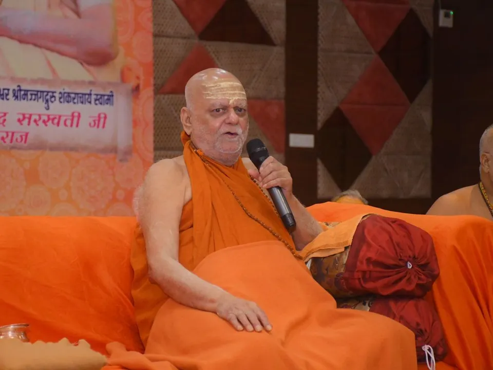 Nishchalananda Saraswati seated on a dais speaking into a handheld microphone, cushions and saffron cloth around him