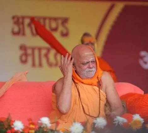 Nishchalananda Saraswati seated on stage with one hand to his head, a banner in Hindi reading Akhand Bharat visible behind him