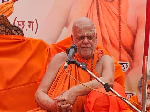 Nishchalananda Saraswati speaking into a microphone at a public programme, a Hindi banner behind him